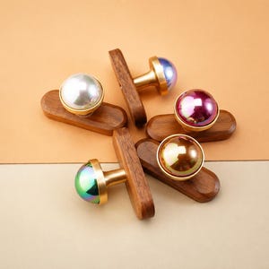 May include: A collection of decorative cabinet knobs. Each knob features a colorful, iridescent, or pearl-like sphere atop a gold-colored base, mounted on a dark wooden rectangular backplate. The knobs are arranged on a two-tone tan and cream background.