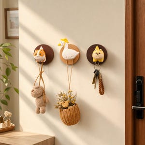 Könnte beinhalten: Drei runde Holzhaken für die Wand mit geschnitzten Tierfiguren: eine Katze, eine Gans mit gelbem Hut und ein Hund. Die Haken halten einen kleinen Plüschbären, einen geflochtenen Korb mit Trockenblumen und einen Schlüsselbund aus Metall.