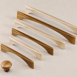 Könnte beinhalten: Eine Sammlung von fünf Metall-Schrankgriffen und einem runden Schrankknopf in abwechselnden gebürsteten Gold- und Bronze-Oberflächen. Jeder Griff hat ein geschwungenes Design mit dem Text No.1Hardware auf der Montagebasis gedruckt.
