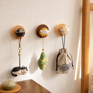 Puede incluir: Tres ganchos de pared circulares de madera montados horizontalmente, cada uno con una pequeña cabeza de animal esculpida. Los ganchos sostienen un dije de gato blanco y negro, un adorno verde con forma de hoja y una bolsa beige.