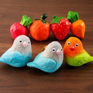 Peut inclure: Ensemble de figurines en bois sculpté à la main représentant des oiseaux et des fruits. Les oiseaux sont peints en bleu, gris, orange et vert. Les fruits comprennent un radis rouge, des tomates oranges, une fraise rouge et des carottes oranges.