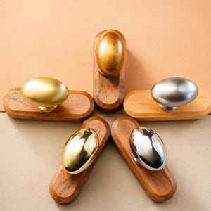 May include: Five oval-shaped cabinet knobs, each mounted on a rectangular wooden base. The knobs feature distinct metallic finishes, including matte gold, polished gold, brushed silver, polished silver, and a warm brass tone, arranged in a star pattern.