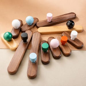 May include: A collection of wooden cabinet pulls with various colored knobs, including white, black, green, pink, blue, and orange. The pulls are made of dark wood and are arranged on a neutral background.