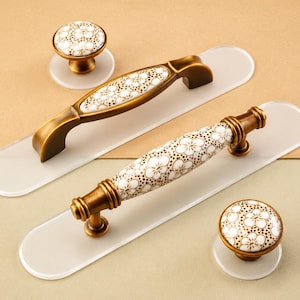 May include: Two brass cabinet pulls and two matching round knobs featuring white ceramic inserts with a delicate brown and gold floral pattern. Each piece is mounted on a clear, oval-shaped acrylic backing plate for installation.