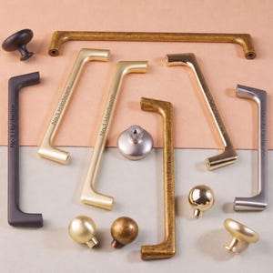Può includere: Una collezione di ferramenta per mobili che include vari pomelli e maniglie per cassetti con finiture in nero opaco, oro spazzolato, ottone antico e argento. Diverse maniglie presentano la scritta No.1 Hardware incisa sul lato.