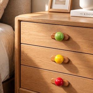 Puede incluir: Una mesita de noche de madera de roble claro con tres cajones, cada uno equipado con una base de mango horizontal de madera y un pomo redondo brillante. El pomo superior es verde lima, el central es amarillo brillante y el inferior es rojo.