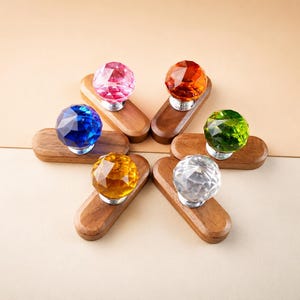 May include: Six drawer knobs featuring faceted, multi-colored crystal spheres in pink, orange, blue, green, yellow, and clear. Each crystal sphere is mounted on a polished silver-tone base attached to an elongated, smooth, dark brown wooden handle.