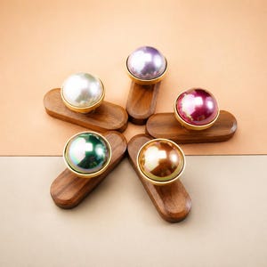 Puede incluir: Cinco pomos decorativos para armarios con esferas nacaradas en blanco, verde, morado, burdeos y dorado, cada uno engastado en un anillo dorado y montado sobre una base de madera oscura. Los pomos están dispuestos en forma de estrella sobre un fondo neutro.