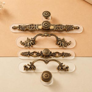 May include: A collection of antique brass-toned metal cabinet hardware, including three drawer pulls with ornate scrollwork and two circular knobs with floral detailing, each mounted on a clear, rounded rectangular acrylic base.