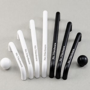 Puede incluir: Una colección de cuatro tiradores de armario blancos y tres negros, cada uno con el texto No.1Hardware impreso en el frente. El conjunto incluye dos pomos esféricos, uno blanco y uno negro, junto a tiradores metálicos curvos.