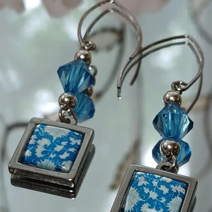 Puede incluir: Un par de pendientes colgantes con ganchos y marcos cuadrados de metal plateado. Cada pendiente contiene una pieza con estampado floral azul y blanco, acentuada por dos cuentas de cristal azul facetado y pequeñas cuentas plateadas.