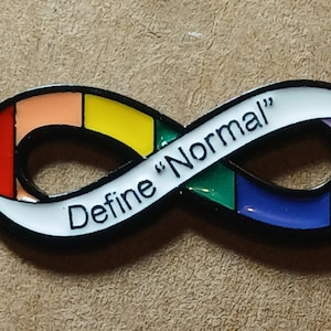 May include: An infinity-shaped enamel pin with a black metal outline. The surface is divided into segments of red, orange, yellow, green, blue, and purple, with a white section featuring the black text "Define "Normal"" printed across it.