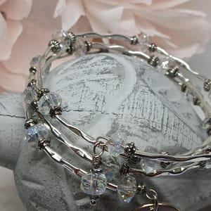 May include: A multi-strand silver-tone wire bracelet featuring a series of clear, faceted crystal beads interspersed with small metallic spacers. The design includes a dangling charm element and a flexible, coiled structure that wraps around.
