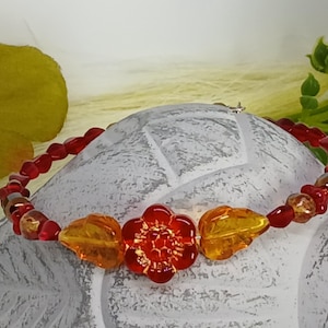 Könnte beinhalten: Ein Perlenarmband mit einer zentralen roten Glasblütenperle, die von zwei bernsteinfarbenen blattförmigen Perlen flankiert wird. Der Rest des Bandes besteht aus kleinen, durchscheinenden roten Perlen und vereinzelten metallischen Akzentperlen.