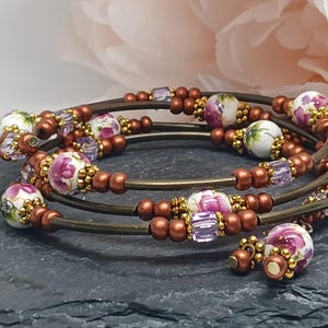 May include: A multi-strand wrap bracelet featuring metallic bronze wire coils, interspersed with round white ceramic beads decorated with pink floral patterns, small metallic gold spacer beads, and translucent light purple square glass beads.