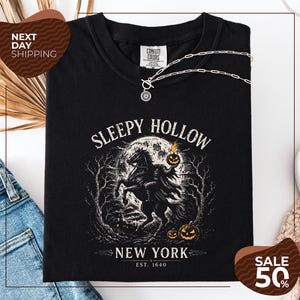May include: A black t-shirt featuring a graphic of the Headless Horseman on a horse against a full moon, with the text SLEEPY HOLLOW NEW YORK EST. 1640. The design includes orange pumpkins, bare trees, and a small house in the background.