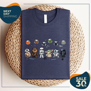 a blue t-shirt with a graphic design of various Star Wars characters, including Chewbacca, Stormtrooper, Baby Yoda, Darth Vader, and others, arranged in a row.