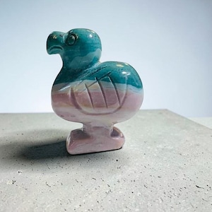 May include: A carved stone dodo bird figurine. The bird has a teal head and upper body, transitioning to pink and white on the lower body and feet. The sculpture is set against a neutral background.