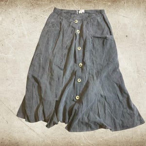 May include: A charcoal gray midi skirt featuring a front button-down closure with light-colored buttons, an elasticized waistband, and two side pockets. The skirt has a flared silhouette and is constructed from a soft, textured fabric.