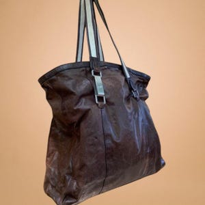 May include: A dark brown leather tote bag with a soft, textured finish. The bag features two long, light-colored fabric shoulder straps attached with silver-toned metal hardware. The design includes a vertical seam down the center of the front panel.