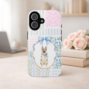 Puede incluir: Una funda para smartphone con un diseño de patchwork de damasco rosa, rayas azules y motivos florales. Un marco ovalado central con un lazo azul claro contiene una ilustración en acuarela de un pastor alemán con un lazo de cuadros azules.