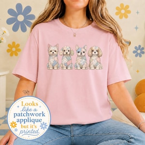 Op de afbeelding: Een lichtroze T-shirt met korte mouwen en een afbeelding van vier honden met patchwork-patronen in blauw, wit en beige. Een ronde badge linksonder toont de tekst: Looks like a patchwork applique but it's printed.