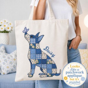 May include: A cream-colored tote bag featuring a printed graphic of a dog composed of blue and white patchwork patterns, including gingham, polka dots, and floral motifs. A blue butterfly rests on the dog's nose, and the name Daisy is printed in blue.
