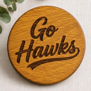 May include: A round wooden coaster with the words "Go Hawks" engraved in a dark brown script. The coaster has a natural wood grain finish and a light brown color. The coaster is a sports-themed item.