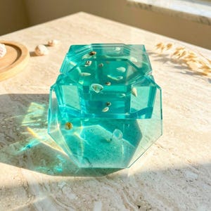 May include: A turquoise resin box with an octagonal shape, embedded with small seashells. The box is translucent, allowing light to pass through, and sits on a marble surface. The box has a lid.