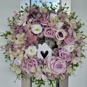 May include: A floral wreath with faux flowers in pink, white, and beige, with green foliage. A white wooden house shape with a heart cutout is at the centre. The wreath is displayed on a mirrored surface. The overall design is soft and inviting.