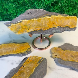May include: A Bumblebee Jasper slab rests on a rose gold metal display stand, accompanied by three additional smaller, irregular slabs of the same stone. Each piece features a distinct horizontal band of vibrant yellow and orange crystalline material.