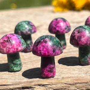 May include: A group of small, mushroom-shaped sculptures. The mushroom caps are a vibrant mix of pink and green, while the stems are green. These decorative items are made of polished stone and are arranged on a wooden surface.