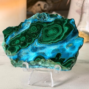 May include: A polished slab of malachite and chrysocolla mineral featuring vibrant bands of deep forest green and bright electric blue. The stone displays natural concentric circular patterns and organic textures, resting on a clear acrylic stand.