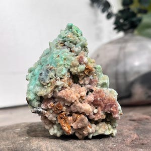 Peut inclure: Gros plan sur un spécimen minéral avec une surface texturée. Le minéral présente un mélange de couleurs, dont le vert clair, le rose et le marron. La forme générale est irrégulière et groupée, suggérant une formation naturelle. Le minéral est exposé sur une surface en pierre.
