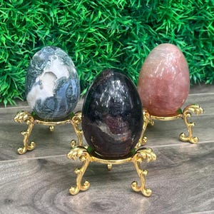 May include: Three polished stone eggs displayed on ornate gold-coloured metal stands. The left egg is mottled grey and white, the centre egg is dark brown with black inclusions, and the right egg is a translucent pale pink with internal veining.