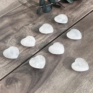 Puede incluir: Ocho piedras pequeñas, lisas y con forma de corazón hechas de cuarzo blanco translúcido. Cada piedra presenta una superficie pulida y una forma redondeada y simétrica. Las piedras están dispuestas de forma dispersa, mostrando su textura cristalina clara y su color blanco pálido constante.