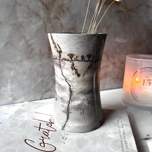 Peut inclure: Un vase cylindrique en pierre présentant une surface lisse gris clair avec des inclusions dendritiques brun foncé naturelles ressemblant à des motifs d'arbres ramifiés. Le vase a un profil légèrement concave et contient quelques tiges séchées.
