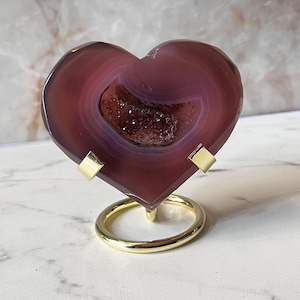 May include: A heart-shaped slice of deep purple agate stone featuring a central druzy crystal cavity. The polished stone is displayed on a gold-toned metal stand with a circular base and two small clips that hold the agate securely in place.