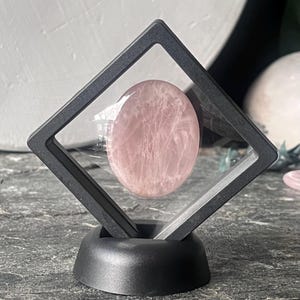 May include: A pink rose quartz gemstone displayed in a black, diamond-shaped frame with a black stand. The gemstone is oval-shaped and has a translucent appearance with visible internal patterns. The display case is clear, allowing a full view of the stone.