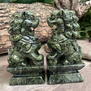 May include: A pair of dark green stone statues depicting guardian lions, each featuring intricate carved manes, open mouths, and detailed muscular bodies, standing on rectangular bases with a polished, marbled texture and deep green color.
