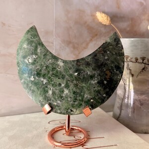 May include: A crescent moon-shaped carving made of dark green, crystalline stone with visible mineral inclusions. The stone is mounted on a polished copper-coloured metal stand featuring a circular base and two small clips holding the piece upright.