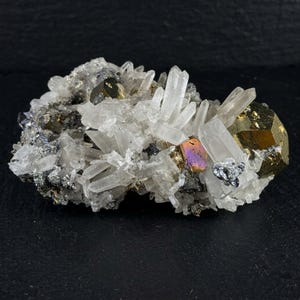 Peut inclure: Un spécimen minéral composé d'un amas de cristaux de quartz transparents et pointus, entremêlés de cubes de pyrite dorés métalliques et de galène gris foncé. Un cristal de pyrite présente un éclat irisé aux reflets violets et orange.