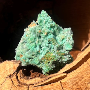Puede incluir: Primer plano de un vibrante espécimen mineral verde claro con una superficie texturizada y cristalina. El mineral tiene áreas de verde más oscuro y parches de marrón anaranjado. El espécimen se muestra sobre una superficie de madera natural.