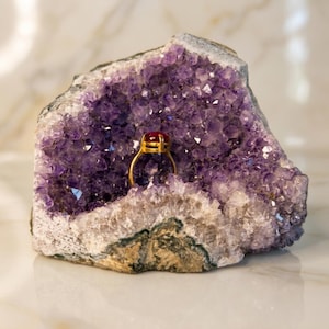 May include: A gold ring featuring a smooth, deep red oval gemstone set in a prong setting. The ring is positioned inside a large, hollowed-out amethyst geode, which displays numerous small, faceted purple crystals throughout its interior cavity.