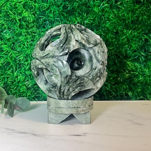 May include: A spherical green stone sculpture featuring intricate, carved openwork patterns that reveal a smaller, solid sphere nested within the centre. The object rests on a matching circular stone base with three small, sturdy feet.