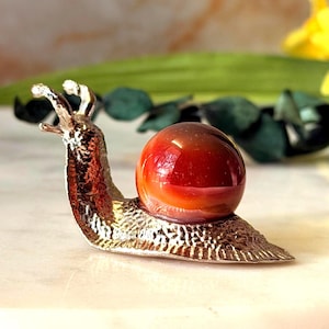 May include: A decorative snail figurine featuring a textured, metallic silver-toned body and a smooth, spherical, deep orange-red gemstone shell. The snail is depicted with two extended eye stalks and a detailed, rippled surface on its base.