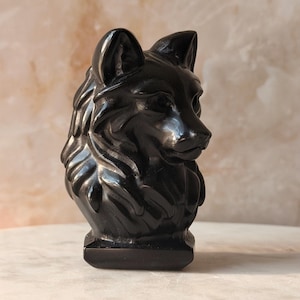 Peut inclure: Une sculpture en pierre noire polie représentant un buste de tête de loup. La pièce présente des textures de fourrure détaillées autour du cou et des oreilles pointues, avec une surface lisse et réfléchissante. Le loup est représenté de profil.