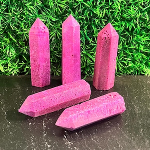 May include: Five vibrant magenta crystal points with a porous, textured surface. Three of the points are standing upright in a vertical position, while two of the points are lying horizontally on a dark, flat surface.