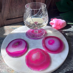 May include: A cut-crystal wine glass with a pink base rests on a matching coaster. Three pink agate coasters are displayed on a white marble surface. A pink tulip adds a splash of colour to the arrangement.