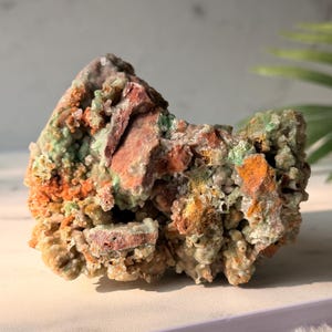 May include: A raw mineral specimen featuring a cluster of crystalline structures in shades of pale green, earthy brown, and vibrant orange. The rock formation displays a rough, jagged texture with varied mineral deposits and small, sparkling crystal points.